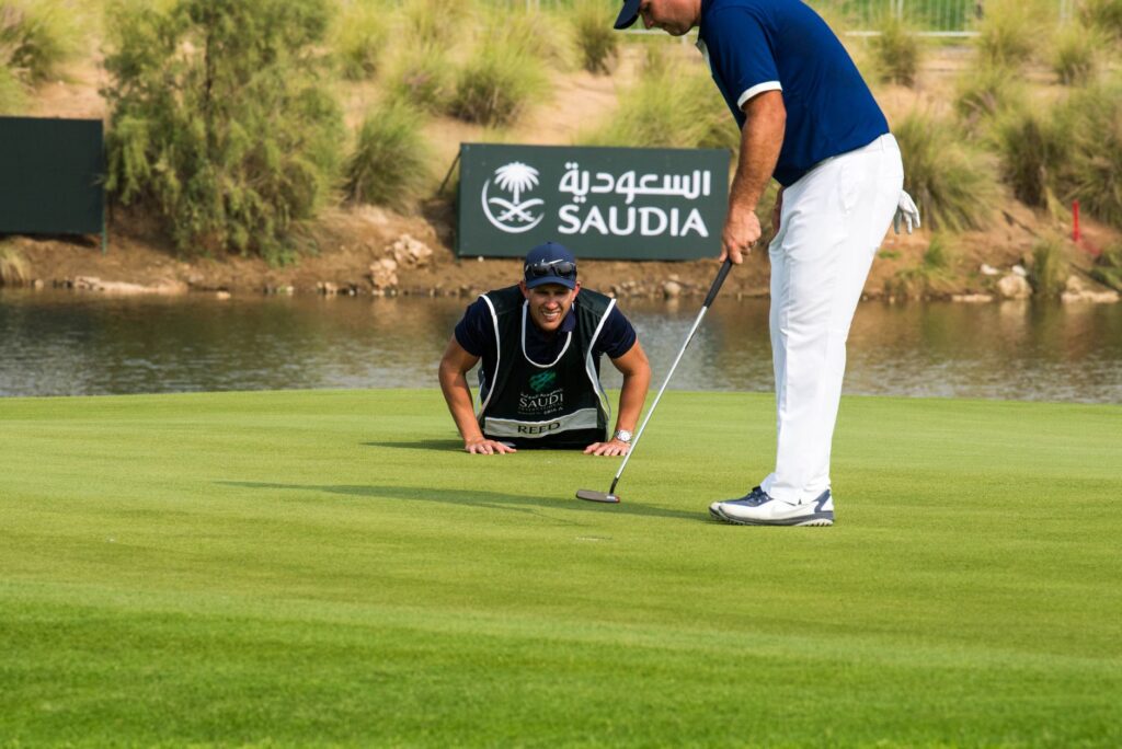 Golfer putting with caddy support at Jeddah, showcasing professional golf equipment and skills.
