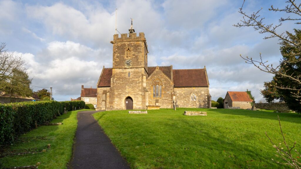 A picturesque stone church with a bell tower in Templecombe, England, surrounded by lush greenery.