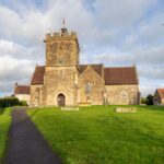 A picturesque stone church with a bell tower in Templecombe, England, surrounded by lush greenery.
