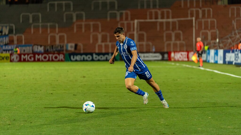 A soccer player in blue jersey dribbling the ball on a well-lit field during a night match.
