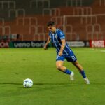 A soccer player in blue jersey dribbling the ball on a well-lit field during a night match.