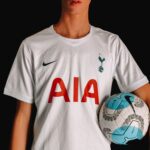 Adult soccer player in Tottenham Hotspur jersey holding a ball against a dark background.