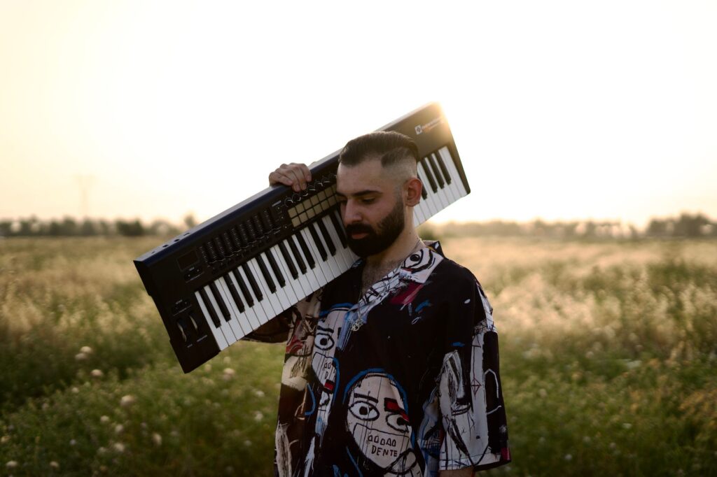 A musician carries a keyboard in a sunlit field in Baghdad, embracing a serene, artistic moment.