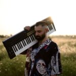 A musician carries a keyboard in a sunlit field in Baghdad, embracing a serene, artistic moment.