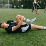 A female soccer player lying on the field, holding her injured leg in pain during a game.