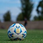 Soccer ball on a grassy field with blurred players in the background, outdoors.