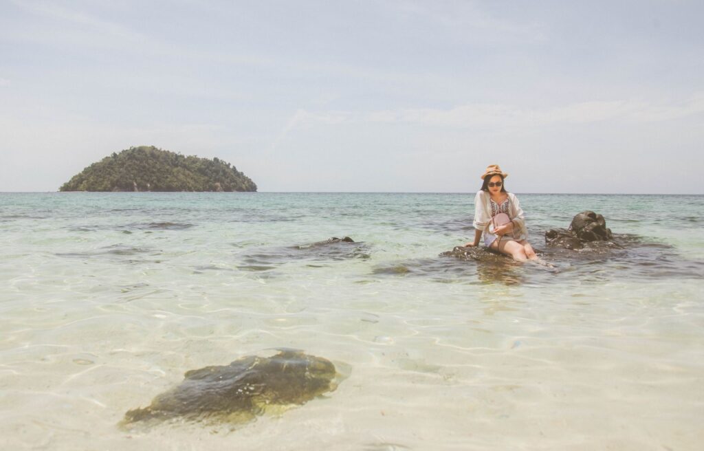 Woman sitting on rocks by the sea with a hat, enjoying a tropical beach view.