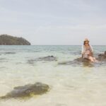 Woman sitting on rocks by the sea with a hat, enjoying a tropical beach view.