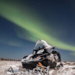 Snowmobile parked in snowy landscape illuminated by Northern Lights at night.