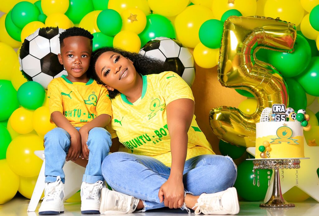 Mother and child in yellow jerseys pose with soccer-themed birthday decorations for a fifth celebration.