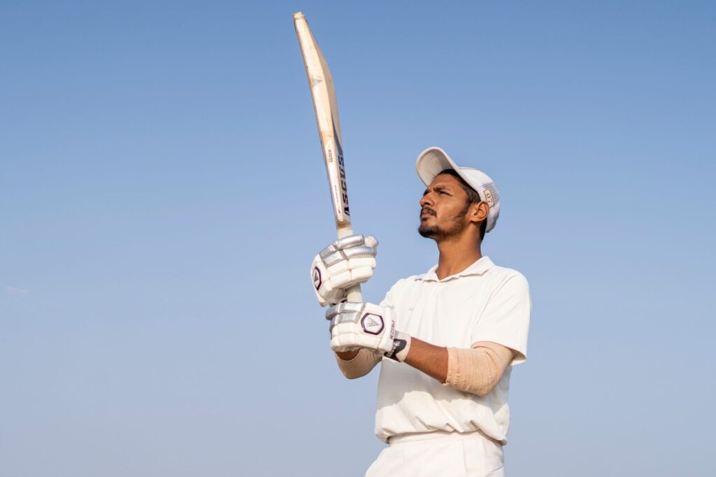 A cricket player in white uniform holds a bat, ready to play under the clear blue sky.