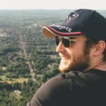 Smiling man in sunglasses and cap enjoying a scenic outdoor view from a hilltop.