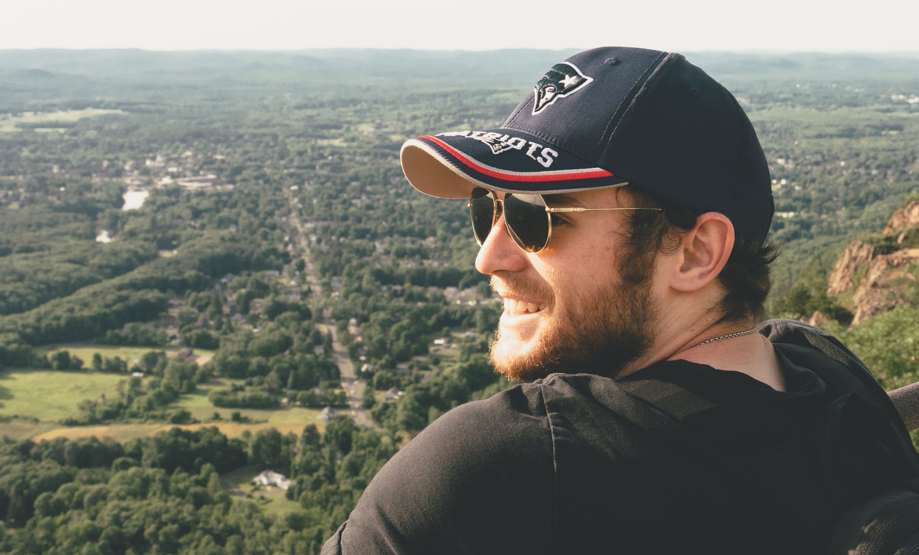 Smiling man in sunglasses and cap enjoying a scenic outdoor view from a hilltop.