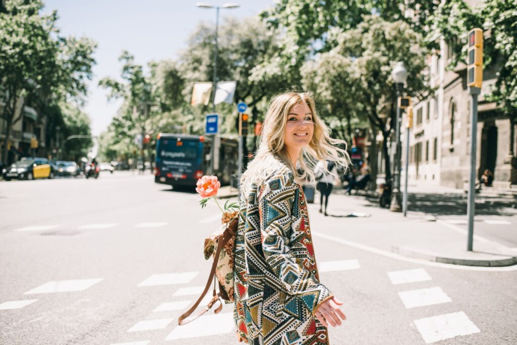 Smiling woman in geometric coat with a flower-adorned backpack crossing a sunny urban street.