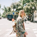 Smiling woman in geometric coat with a flower-adorned backpack crossing a sunny urban street.