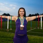 Smiling female athlete holding numerous medals on an outdoor sports field.