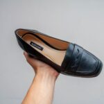 Close-up of a hand holding a black leather shoe by Alex Marie against a plain background.