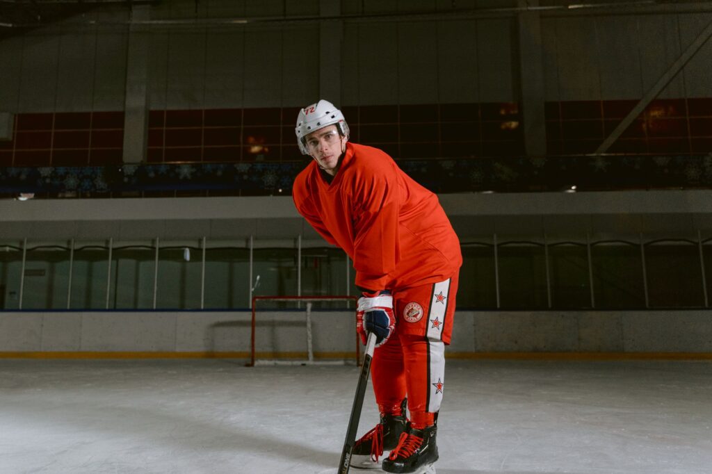 Male hockey player in red uniform and helmet posing on an indoor ice rink.