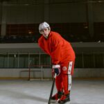 Male hockey player in red uniform and helmet posing on an indoor ice rink.