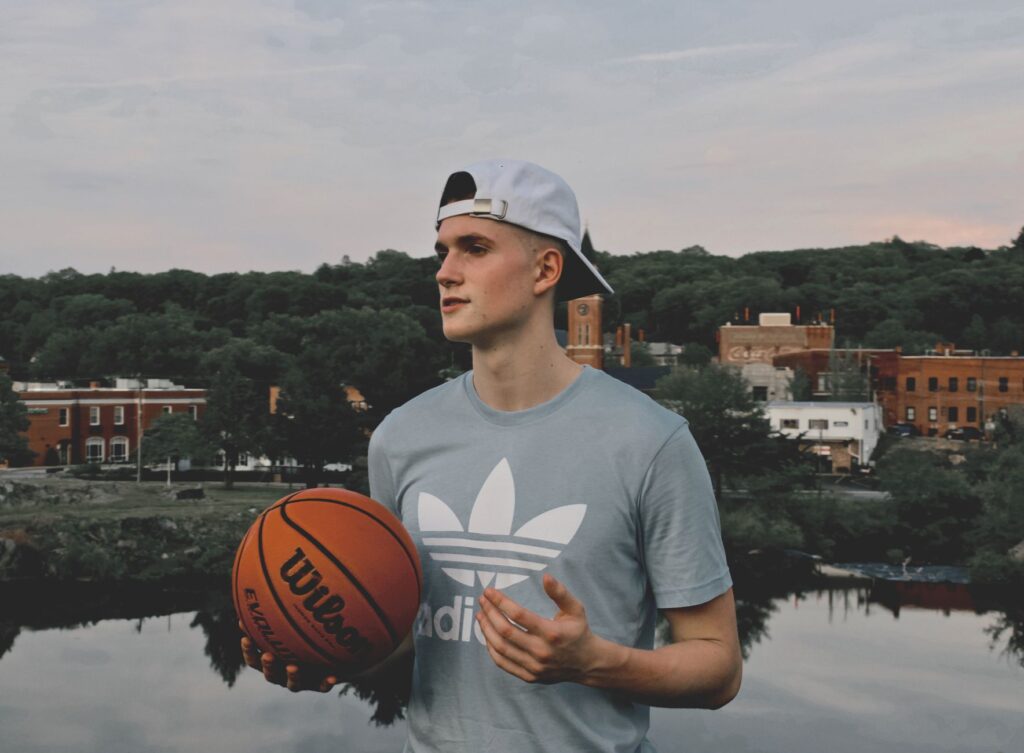 Young man holding a basketball by a lake in Putnam, CT, with town buildings in the background.