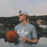 Young man holding a basketball by a lake in Putnam, CT, with town buildings in the background.