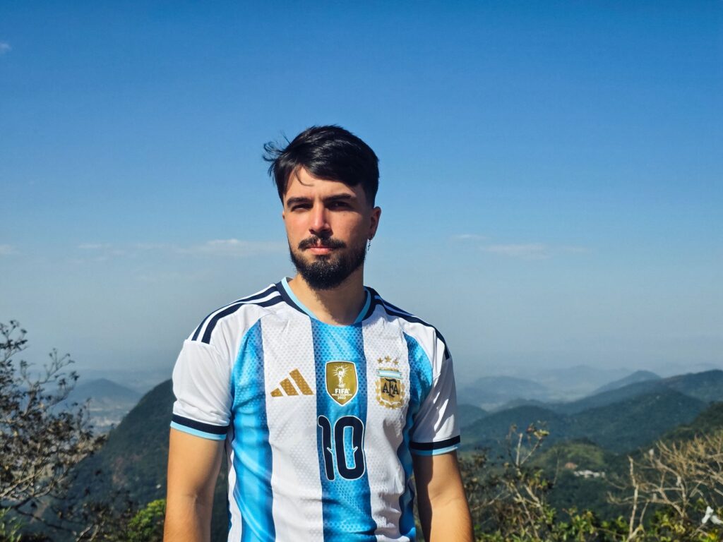 A man in an Argentina football jersey stands with a scenic mountain backdrop under a clear sky.
