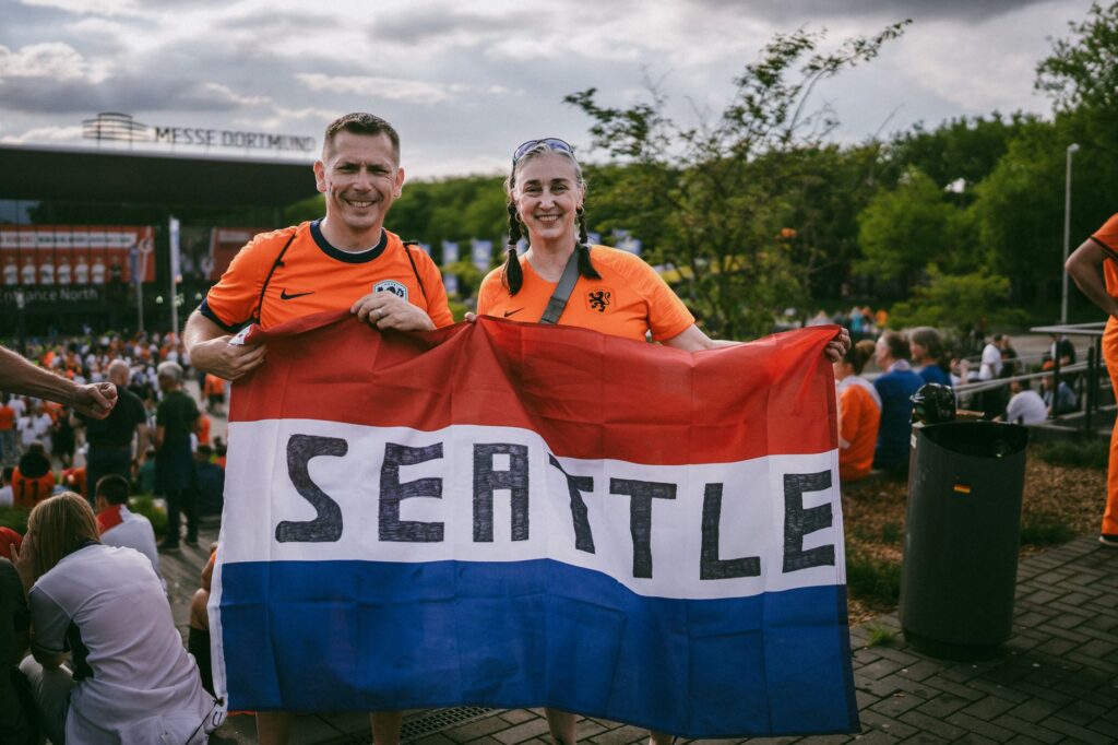 Two soccer fans in orange jerseys holding a Seattle flag outside a stadium in Dortmund.