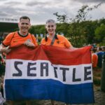 Two soccer fans in orange jerseys holding a Seattle flag outside a stadium in Dortmund.