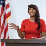 Black woman giving a speech at a podium with an American flag in the background.
