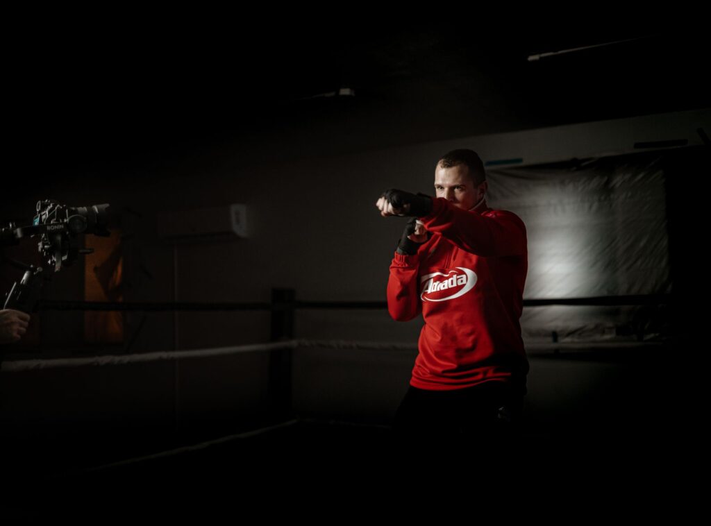 Powerful male athlete in sportswear practicing boxing hits in ring against crop anonymous partner recording video on photo camera