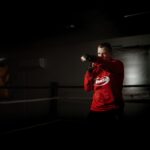 Powerful male athlete in sportswear practicing boxing hits in ring against crop anonymous partner recording video on photo camera