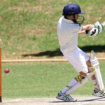 Young cricket player in full gear performing a sweep shot on a sunny day outdoors.