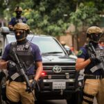 SWAT team members in tactical gear standing by a police vehicle during an operation. Outdoors urban setting.