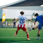 Two teenage boys playing soccer on a green field, mid-action kick under bright daylight.