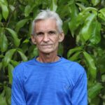 A senior man in a blue shirt stands outdoors against a lush green foliage background. Outdoor portrait.