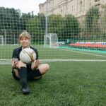 A female soccer player sitting with a ball on a soccer field, enjoying a break.