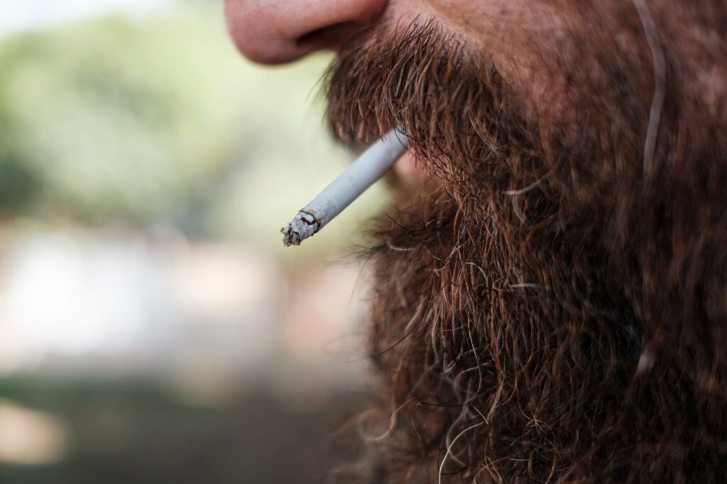 A detailed close-up of a bearded man smoking a cigarette outdoors, showcasing lifestyle choices.