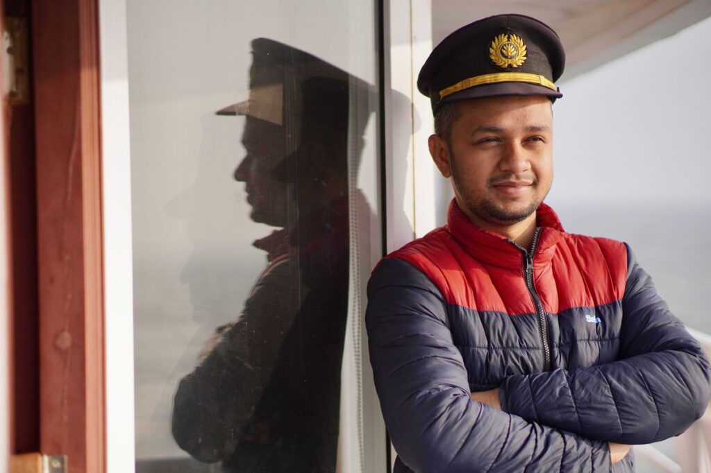 A ship officer in uniform reflects on glass, displaying confidence against a serene sea backdrop.