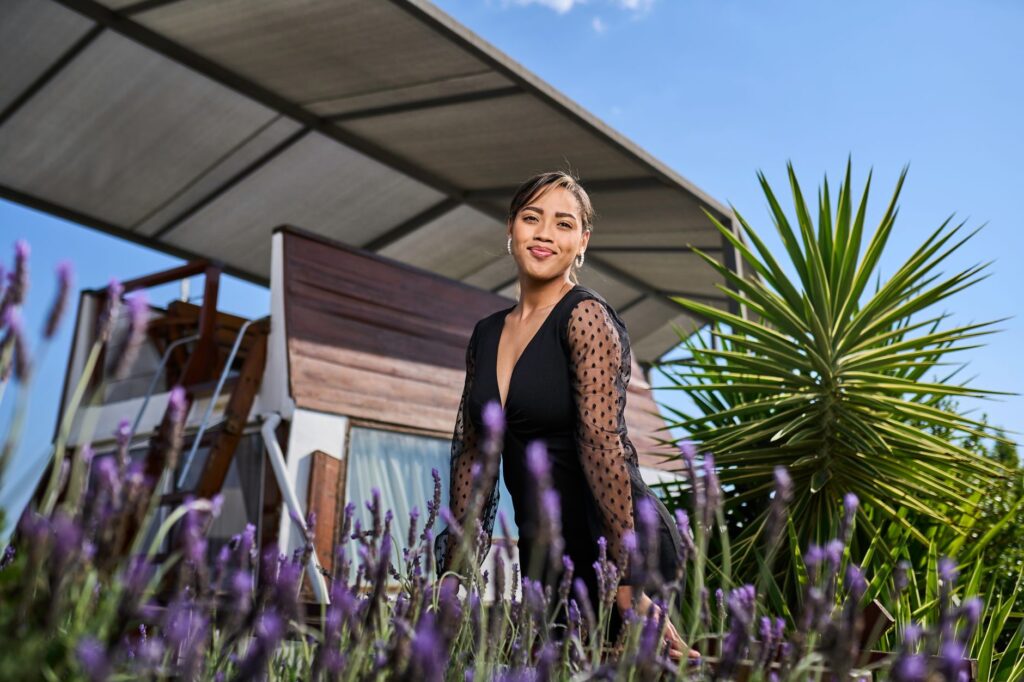 A smiling woman in a stylish black dress poses outdoors amidst lavender flowers and a modern structure.