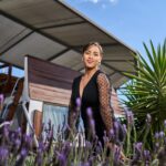 A smiling woman in a stylish black dress poses outdoors amidst lavender flowers and a modern structure.
