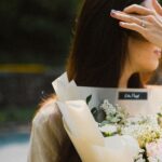 A woman with long hair holds a bouquet of soft-colored flowers outdoors, creating an elegant and serene atmosphere.