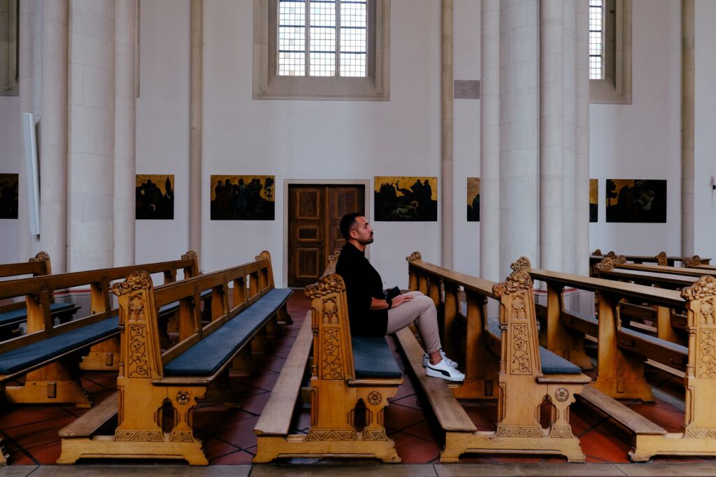 A man sits alone in a church pew, surrounded by ornate wooden benches and religious art.