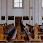 A man sits alone in a church pew, surrounded by ornate wooden benches and religious art.