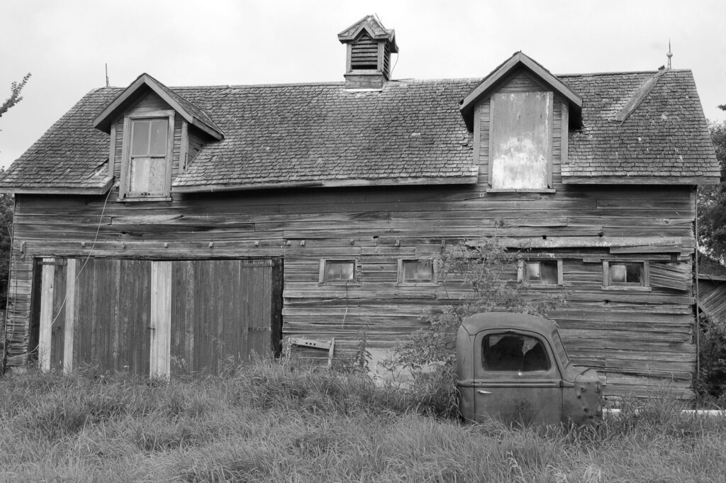 Black and white photo of an old barn with a rusty truck in front, evoking rustic nostalgia.