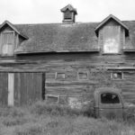 Black and white photo of an old barn with a rusty truck in front, evoking rustic nostalgia.