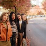 Group of stylish women posing on a tree-lined city street in autumn.