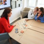 Three women playing a card game while sitting at a table in an office environment.