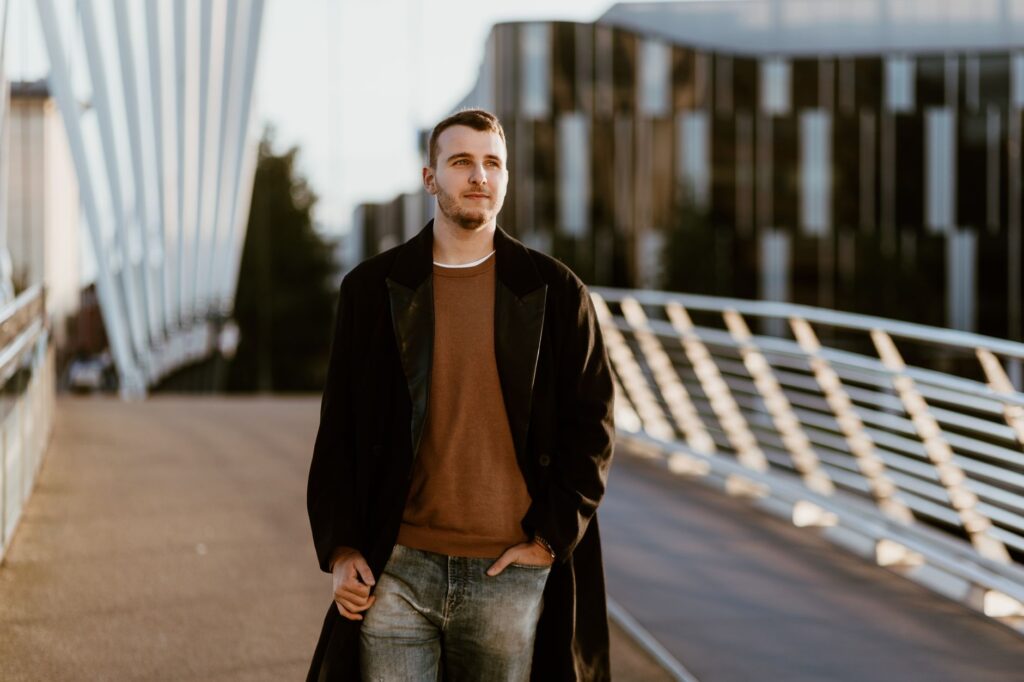 Young man in fashion coat walking on a modern bridge in Manchester, capturing urban style.
