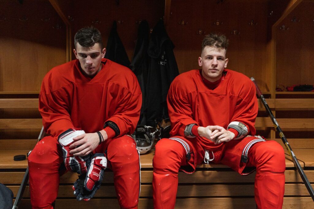 Two hockey players in red gear sit in a locker room resting before or after a game.