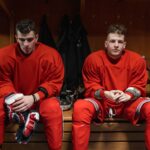 Two hockey players in red gear sit in a locker room resting before or after a game.